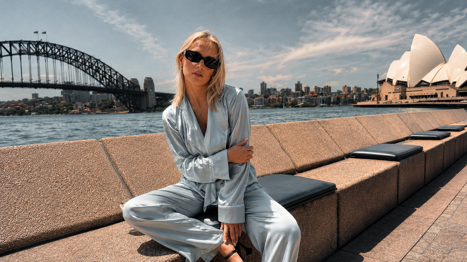Woman in a light outfit sitting on a bench with the Sydney Harbour Bridge and Opera House in the background.